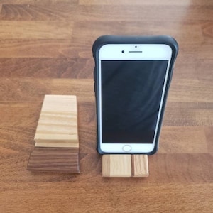 May include: A wooden phone stand with a light brown top and a dark brown bottom. A black smartphone is resting on the stand.