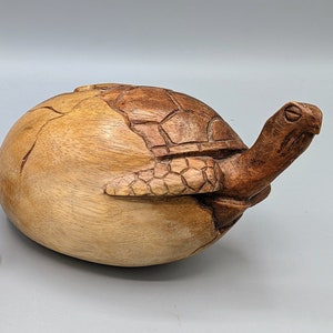 Może przedstawiać: Drewniana rzeźba żółwia z brązowym i beżowym wykończeniem. Żółw jest rzeźbiony w realistycznym stylu i ma szczegółowy pancerz. Rzeźba stoi na jasnoszarym tle.