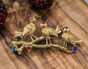 May include: Gold-tone brooch featuring three birds perched on a branch with colorful rhinestones. The birds have red eyes and the branch has green, blue, and red rhinestones.