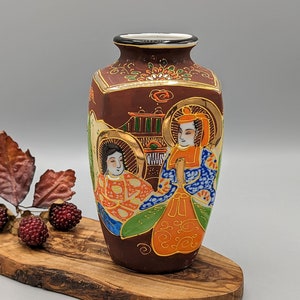 May include: A small, brown ceramic vase with gold accents and a hand-painted scene of two figures in traditional Japanese clothing. The vase is decorated with flowers and geometric patterns.