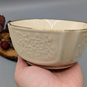 May include: A white porcelain bowl with a gold rim and embossed floral design. The bowl is slightly angled and has a scalloped edge.