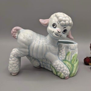 May include: A ceramic planter shaped like a light blue lamb with pink accents. The lamb is standing on its hind legs with its front legs resting on a green and brown planter base.
