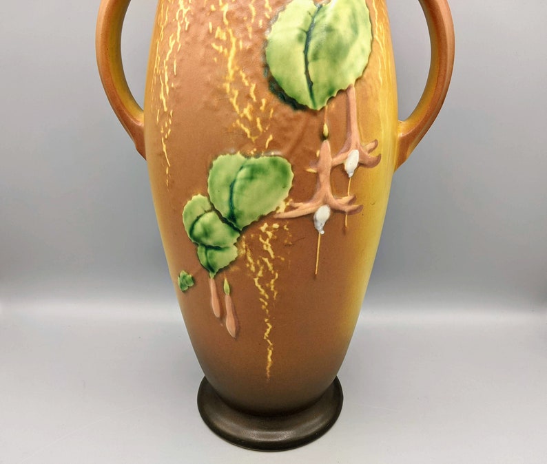 Może przedstawiać: Brązowy ceramiczny wazon z dwoma uchwytami i wzorem kwiatowym. Wazon ma fakturowaną powierzchnię i brązową glazurę. Wz&oacute;r kwiatowy obejmuje zielone liście i r&oacute;żowe kwiaty.