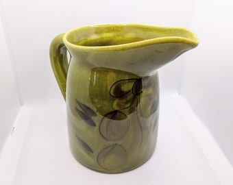 May include: A green ceramic pitcher with a floral design in brown and black. The pitcher has a wide spout and a handle.