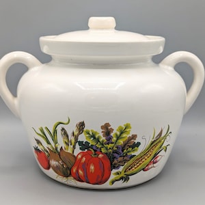 Puede incluir: Cazuela de cerámica blanca con tapa y dos asas. El plato está decorado con una colorida ilustración de varias verduras, como tomates, cebollas y maíz. La tapa tiene un pequeño pomo.