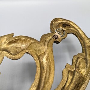 May include: Close-up of a decorative, ornate, gold-coloured metal wall hanging. The design features a flowing, organic shape with intricate details. A small metal loop and screw are visible for hanging.