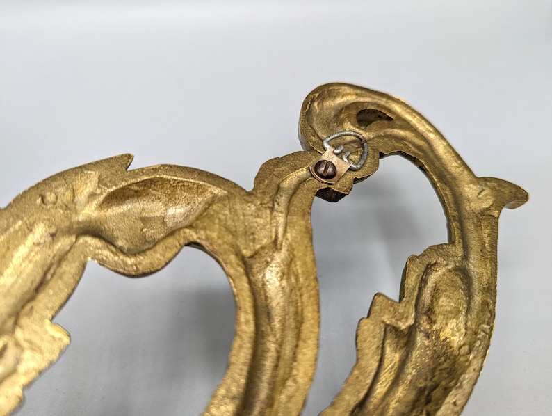 May include: Close-up of a decorative, ornate, gold-coloured metal wall hanging. The design features a flowing, organic shape with intricate details. A small metal loop and screw are visible for hanging.