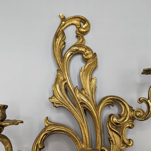 May include: An ornate gold wall sconce with two candle holders. The sconce has a detailed, swirling leaf and floral design. The candle holders are shaped like small cups with a floral base. A decorative piece for home decor.