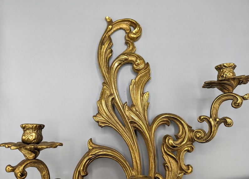 May include: An ornate gold wall sconce with two candle holders. The sconce has a detailed, swirling leaf and floral design. The candle holders are shaped like small cups with a floral base. A decorative piece for home decor.