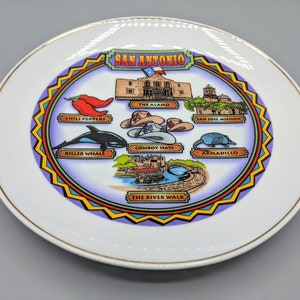 Może przedstawiać: Biały ceramiczny talerz ze złotą obwódką przedstawia kolorową ilustrację zabytków i ikon San Antonio. Talerz zawiera obrazy Alamo, Misji San Jose, papryczek chili, kowbojskich kapeluszy, orki, pancernika i The River Walk.