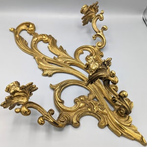 May include: An ornate, gold-coloured wall sconce with a detailed, scrolling leaf and floral design. The sconce features two curved arms with decorative elements and a central flourish. The overall aesthetic is vintage and decorative.