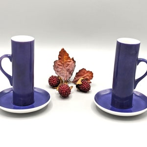 May include: Two indigo-colored coffee mugs with handles, resting on matching saucers with white rims. The mugs are cylindrical and stand upright. A dried leaf and several small, red berries are arranged between the mugs.