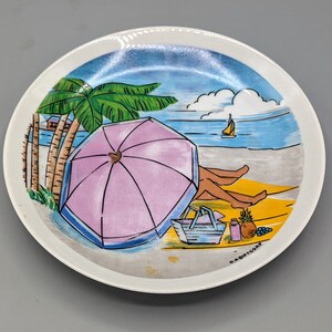 May include: Round ceramic plate with a beach scene. The plate features a pink umbrella, palm trees, and a person relaxing on the beach. A sailboat is in the distance. The plate has a white rim and a painted design.