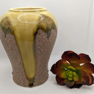 May include: A decorative ceramic vase with a speckled brown base and a light yellow top. The vase has a unique shape and is accented with dark green and brown drips. A faux succulent plant with red, green, and yellow leaves sits beside the vase.