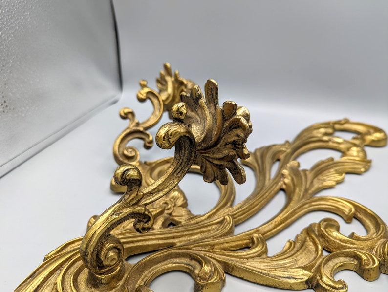 May include: A pair of gold-coloured, ornate wall decorations with a scrolling, floral design. The decorative pieces feature detailed leaf and vine patterns, creating an elegant, vintage aesthetic. The metalwork has a slightly aged appearance, adding to its antique charm.