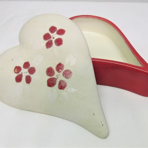 Può includere: Una scatola portaoggetti in ceramica bianca a forma di cuore con un coperchio rosso. La scatola è decorata con tre fiori rossi.
