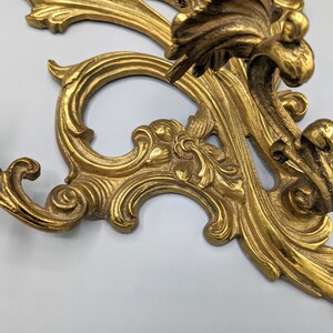 May include: An ornate, gold-coloured metal wall sconce with a detailed, scrolling design. The sconce features a classic, vintage aesthetic with intricate floral and leaf motifs. The overall design is symmetrical and elegant.