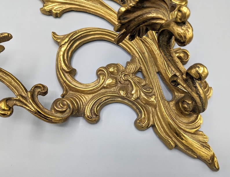 May include: An ornate, gold-coloured metal wall sconce with a detailed, scrolling design. The sconce features a classic, vintage aesthetic with intricate floral and leaf motifs. The overall design is symmetrical and elegant.