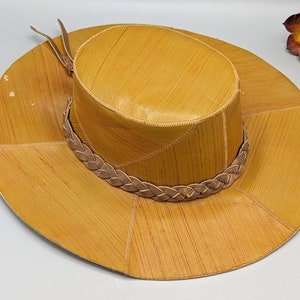 Peut inclure: Un chapeau en cuir brun à larges bords avec une bande en cuir tressée et une sangle en cuir brun. Le chapeau est fait de panneaux de cuir cousus et a une couronne légèrement arrondie.