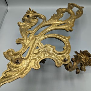 May include: An ornate, gold-coloured metal decorative wall sconce with a scrolling, floral design. The sconce features a detailed, symmetrical pattern with multiple mounting holes. The design is reminiscent of antique decor.