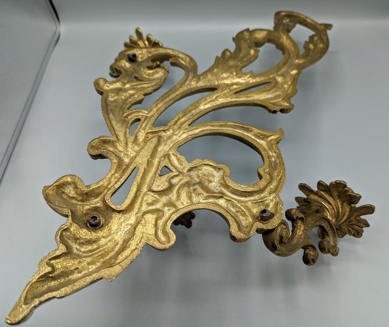 May include: An ornate, gold-coloured metal decorative wall sconce with a scrolling, floral design. The sconce features a detailed, symmetrical pattern with multiple mounting holes. The design is reminiscent of antique decor.