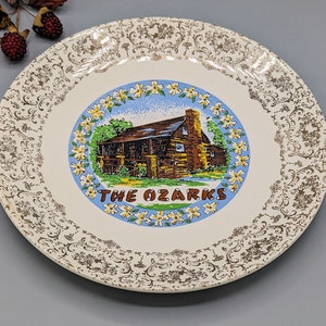 May include: A white ceramic plate with a gold and brown floral pattern. The centre of the plate features a colourful illustration of a cabin in the woods with the text "The Ozarks".