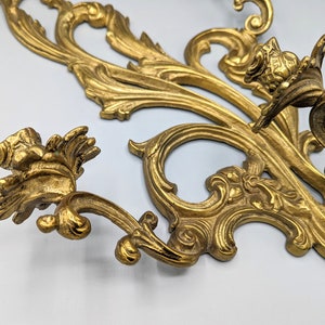 May include: An ornate, gold-coloured wall decoration in an antique style, featuring intricate floral and scrollwork designs. The decorative piece has a detailed, three-dimensional relief with a vintage aesthetic. The metalwork has a slightly aged appearance.