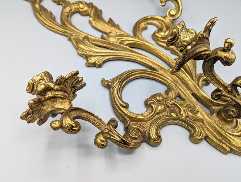 May include: An ornate, gold-coloured wall decoration in an antique style, featuring intricate floral and scrollwork designs. The decorative piece has a detailed, three-dimensional relief with a vintage aesthetic. The metalwork has a slightly aged appearance.