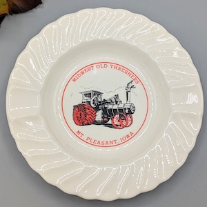 Può includere: Posacenere in ceramica bianca con un'immagine rossa e nera di una macchina a vapore. L'immagine è circondata da un cerchio rosso con il testo "Midwest Old Threshers Mt. Pleasant, Iowa".