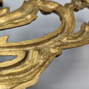 May include: Close-up of a gold-coloured, ornate metal decorative element. The design features a flowing, leaf-like pattern with intricate details and a slightly aged appearance. The element appears to be part of a larger decorative piece.