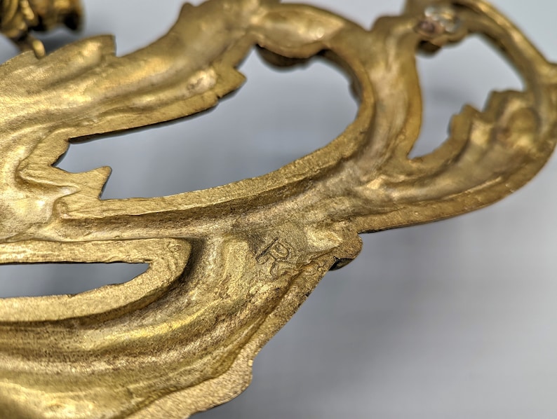 May include: Close-up of a gold-coloured, ornate metal decorative element. The design features a flowing, leaf-like pattern with intricate details and a slightly aged appearance. The element appears to be part of a larger decorative piece.