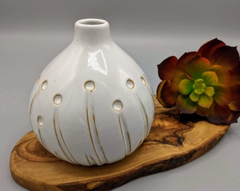 May include: White ceramic vase with a round, bulbous shape and a textured surface. The vase has a gold-colored design of small circles and lines. The vase is sitting on a wooden surface.
