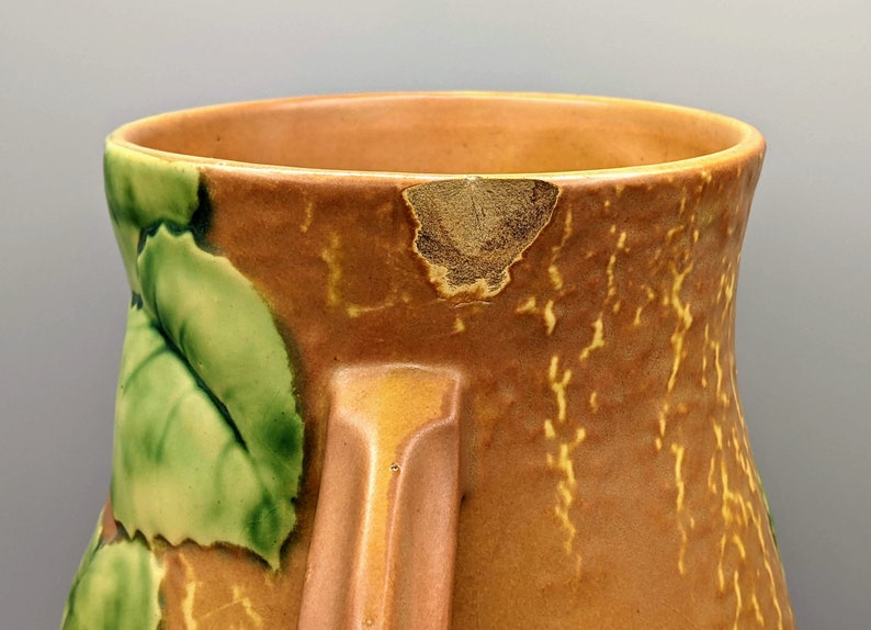 Może przedstawiać: Brązowy ceramiczny wazon z zielonym wzorem liści. Wazon ma odprysk na brzegu i fakturowaną powierzchnię.
