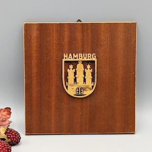 May include: A wooden plaque with a gold-colored cutout of the Hamburg coat of arms. The coat of arms features a castle with three towers and the word "HAMBURG" above it.