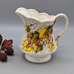Puede incluir: Jarra de cerámica blanca con un diseño en relieve de hojas de otoño en tonos amarillos, naranjas y marrones. La jarra tiene un borde festoneado y un asa.