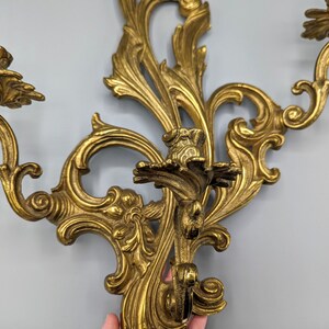 May include: Ornate gold-coloured wall sconce with a floral and scroll design. The sconce has three candle holders and is made of metal. The decorative piece is suitable for a vintage or antique-themed interior.