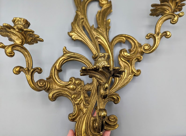 May include: Ornate gold-coloured wall sconce with a floral and scroll design. The sconce has three candle holders and is made of metal. The decorative piece is suitable for a vintage or antique-themed interior.