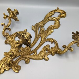 May include: Ornate gold wall sconce with two candle holders. The sconce features a detailed, scrolling design with floral accents. The dimensions are approximately 46 cm wide and 30 cm tall.