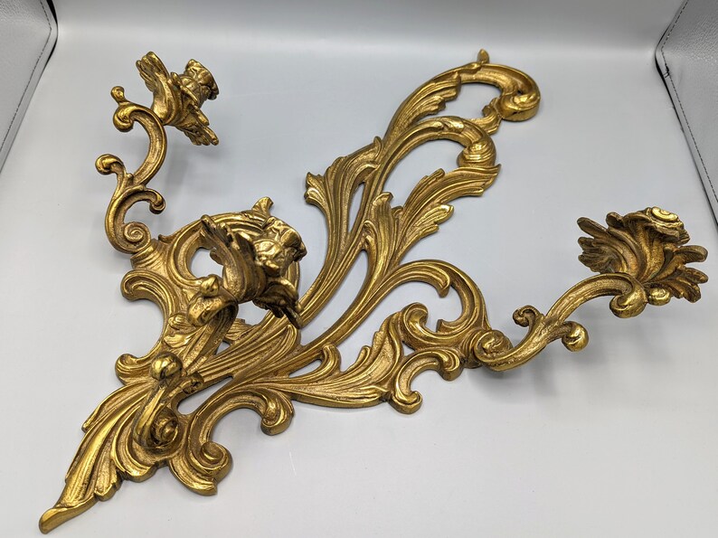 May include: Ornate gold wall sconce with two candle holders. The sconce features a detailed, scrolling design with floral accents. The dimensions are approximately 46 cm wide and 30 cm tall.