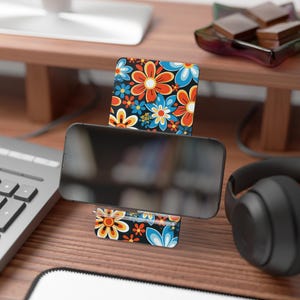 Può includere: Uno smartphone nero è appoggiato su un supporto per telefono con motivo floreale. Il supporto presenta uno sfondo nero con fiori arancioni, blu e bianchi. Il supporto è su una scrivania di legno, con una tastiera e delle cuffie sullo sfondo.