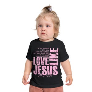 Op de afbeelding: Een zwart t-shirt met korte mouwen en roze verweerde tekst met de tekst A new command I give you: Love one another. As I have loved you, so you must love one another. LOVE LIKE JESUS John 13:34 in een gedurfd, gelaagd grafisch ontwerp.