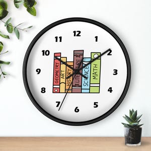 May include: A round wall clock with a black frame and a white face featuring black numbers from 1 to 12. In the centre, a colourful illustration shows a row of books labelled Geometry, Art, History, Literature, Science, and Math with black hands.