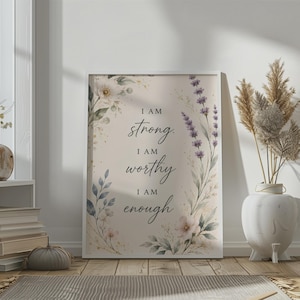 May include: A rectangular framed wall art print featuring a beige background with watercolor floral illustrations of lavender and white flowers. The text in the center reads: I AM strong. I AM worthy. I AM enough. The print is displayed in a white frame.