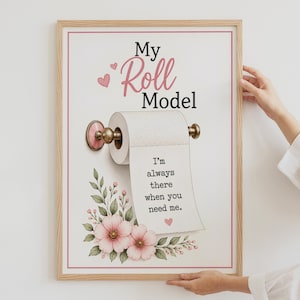 May include: A framed wall art print featuring a toilet paper roll on a pink and gold holder. The text reads My Roll Model and I'm always there when you need me. The design includes pink flowers, green leaves, and small pink hearts on a white background.