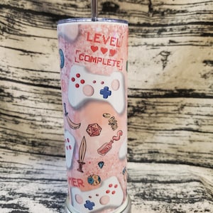 May include: A pink and white tumbler with a gamer theme. It features a video game controller, dice, and the text "Level Complete".
