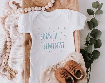 May include: White baby onesie with the text "Born a Feminist" in blue and pink lettering with confetti around the text.