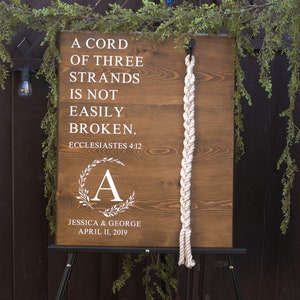 May include: A wooden wedding sign with the text "A CORD OF THREE STRANDS IS NOT EASILY BROKEN." and the names Jessica & George, April 11, 2019. The sign has a white rope detail and a floral wreath with the letter "A".