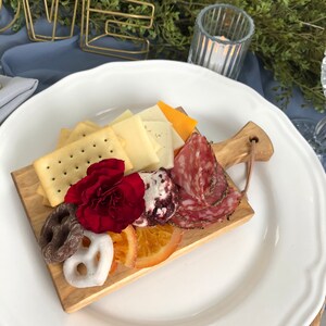 May include: A wooden charcuterie board with crackers, cheese, salami, pretzels, dried orange slices, and a red flower, arranged on a white plate. The board is set on a blue tablecloth, with a gold "LOVE" sign in the background.