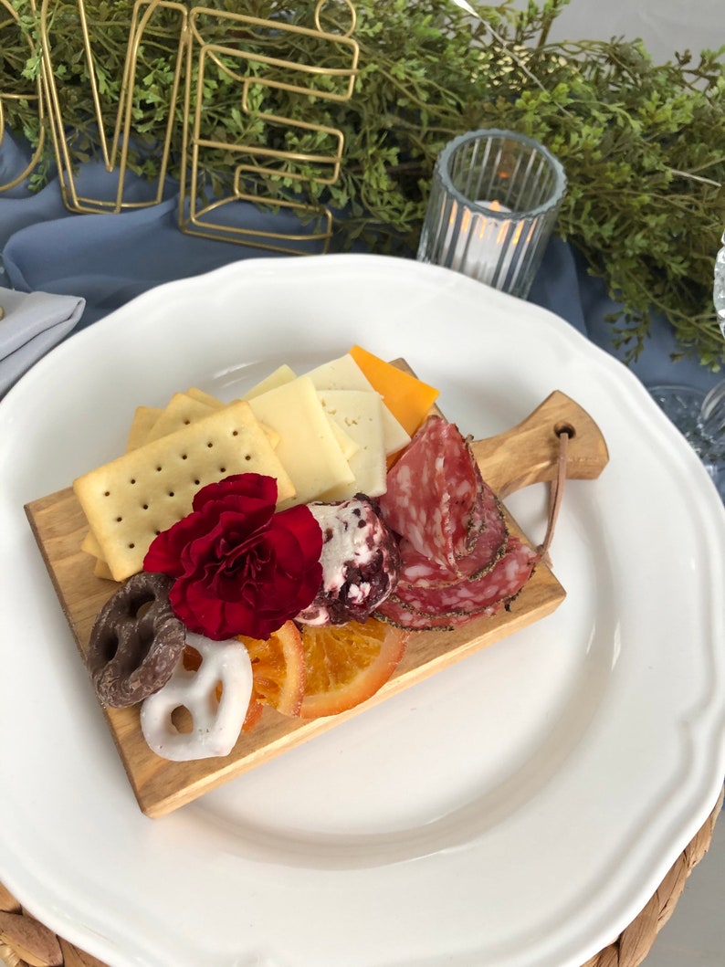 May include: A wooden charcuterie board with crackers, cheese, salami, pretzels, dried orange slices, and a red flower, arranged on a white plate. The board is set on a blue tablecloth, with a gold "LOVE" sign in the background.