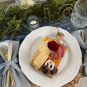May include: A charcuterie board with crackers, cheese, salami, and pretzels on a wooden cutting board. The board is on a white plate with a scalloped edge. Gold silverware and a blue napkin are on the left. The word "love" is in gold wire.